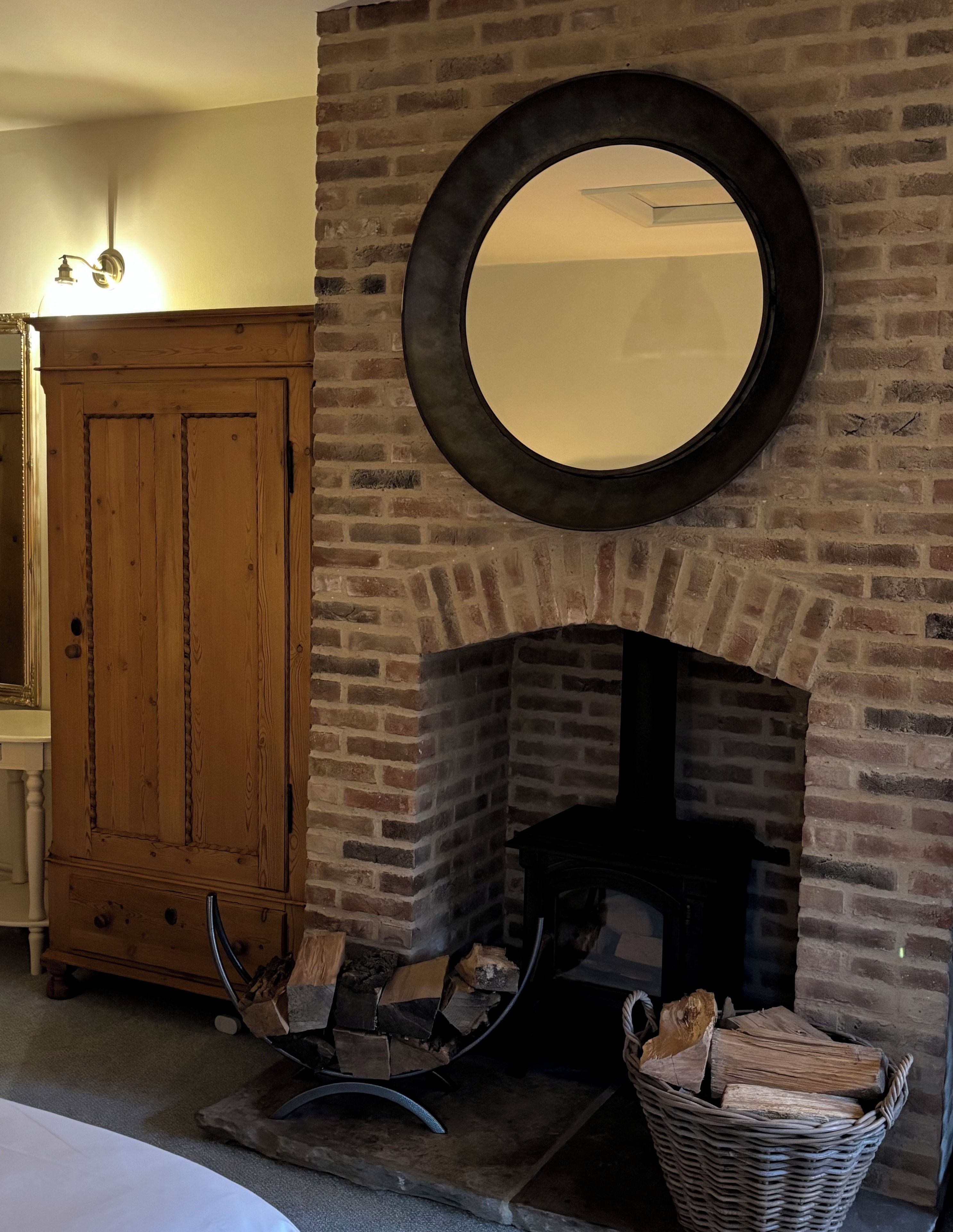 Une cheminée en briques surmontée d’un miroir rond, un meuble en bois à gauche et un panier de bois de chauffage devant.