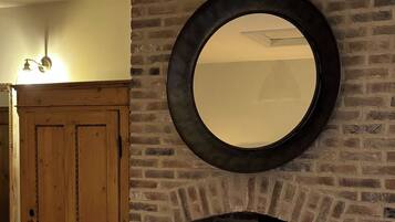 Une cheminée en briques surmontée d’un miroir rond, un meuble en bois à gauche et un panier de bois de chauffage devant.