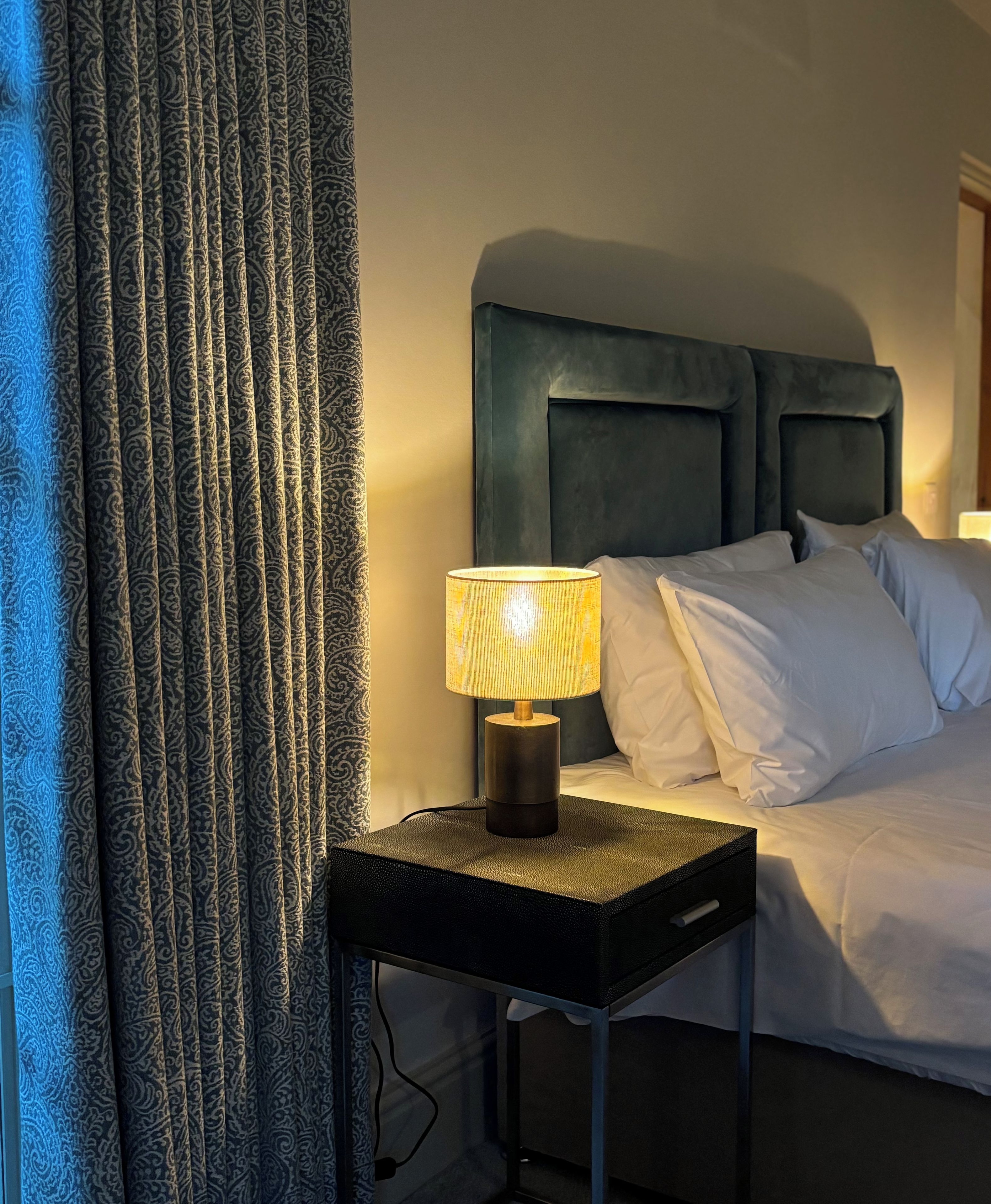 Uma cama com roupa de cama branca, uma cabeceira escura e uma luminária sobre uma mesinha ao lado.