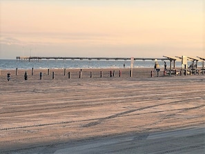 A beach with a pier extending into the water, beach umbrellas, and a few people. - BC9--Studio Sleeps 2 Adults/2 Kids, Shared Pool/Hot Tub, Walk to Beach, Dogs OK! (Port Aransas)