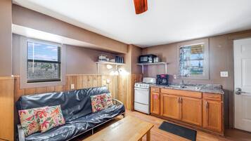 A compact living space with a kitchenette, a sofa with floral cushions, a wooden table, and a view through a window.