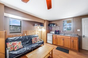 Ein kompakter Wohnraum mit Küchenzeile, einer Couch mit floralen Kissen, einem Holztisch und Blick durch ein Fenster.