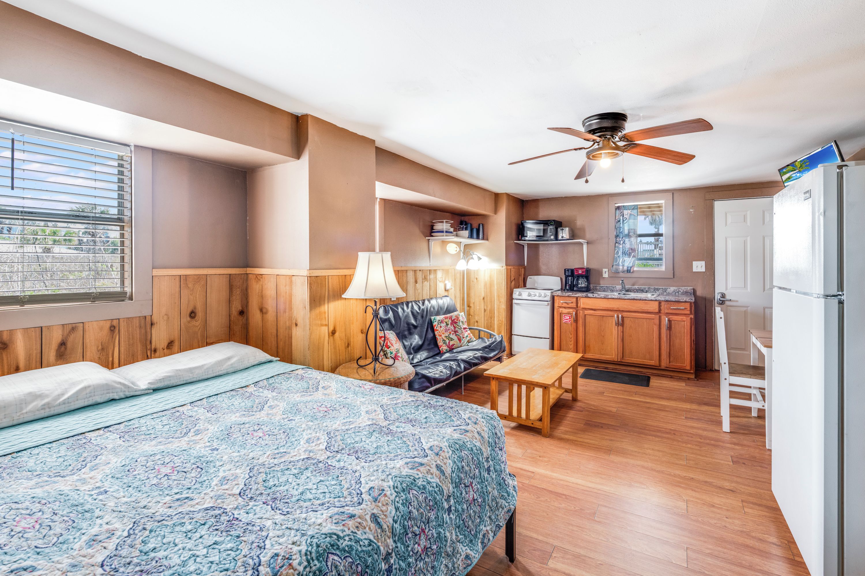 A compact room with a bed, a sofa, a small table, a ceiling fan, a kitchenette, and a window with blinds.