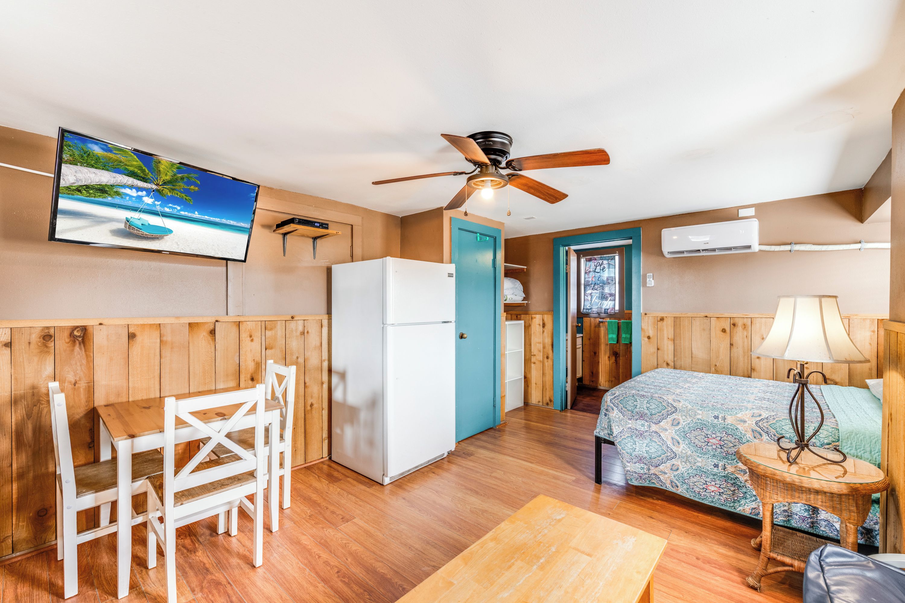 A compact room with a wooden floor, a bed with a patterned bedspread, a white refrigerator, a wooden dining table with chairs, a ceiling fan, and a television displaying a beach scene.