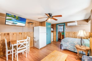 Ein gemütlicher Raum mit Holzboden, einem Bett mit gemustertem Überwurf, einem weißen Kühlschrank, einem Holztisch mit Stühlen, einem Deckenventilator und einem Fernseher, der eine Strandansicht zeigt.