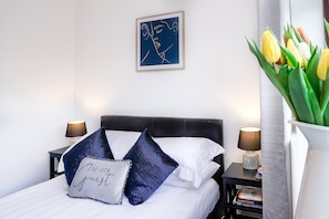 A neatly made bed with white and blue pillows, a framed artwork above it, and a vase with yellow tulips on a bedside table. - 2-bed Cambridge house with garden and parking for 2 cars (Fulbourn)