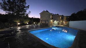 A swimming pool with stone tiles, lounge chairs, and a stone building with lights. - Studio 10 (Kato Platres)