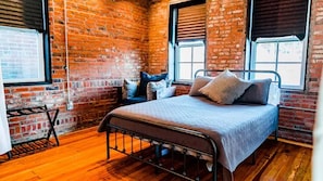 A room with a brick wall, a bed with pillows, a nightstand, and a window. - Beautiful 4-Bed Loft in the heart of Festus Main St (Festus)