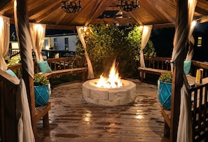 En udendørs terrasse med en rund ildsted i sten, træterrasse og siddepladser.
