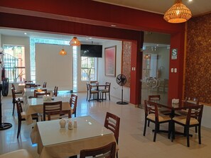 Ein Restaurant mit mehreren Tischen und Stühlen, einem an der Wand befestigten Fernseher und einem Stehlüfter.