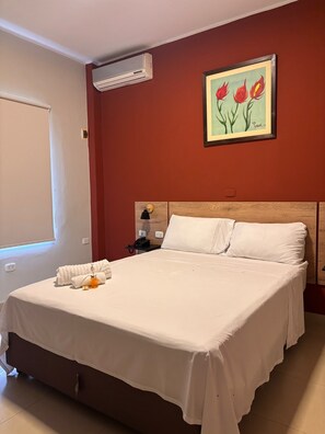 A hotel room with a bed, a nightstand, an air conditioning unit, a painting, and a window.