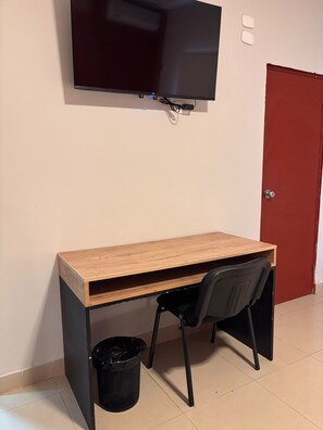 A small, enclosed room with a wooden desk, a black chair, a black trash bin, and a television mounted on the wall.