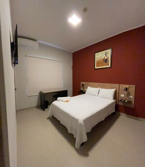 A hotel room with a bed, a desk, a chair, a TV mounted on the wall, and a painting on the wall.