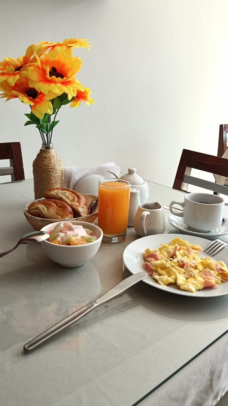 Desayuno con huevos revueltos, un bol de fruta, un vaso de zumo de naranja, un florero y una cesta de pan sobre una mesa.