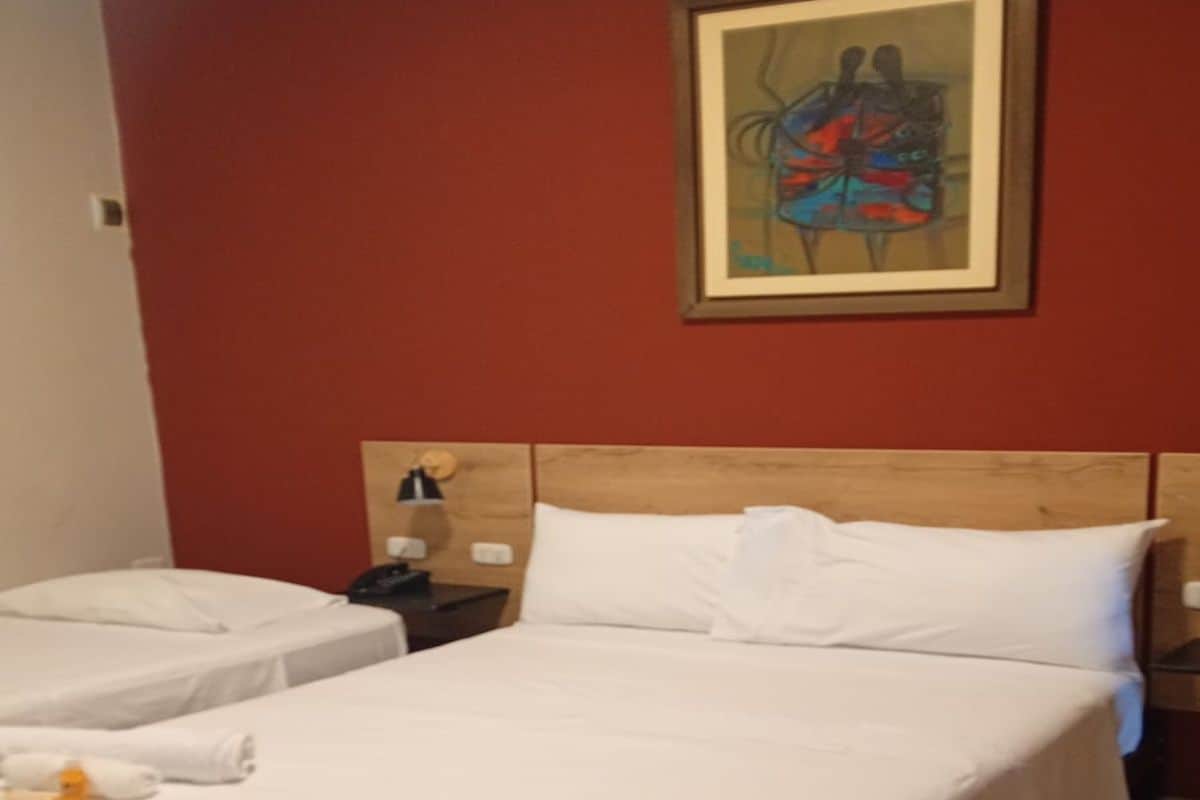 A hotel room with a double bed, a single bed, a nightstand with a lamp, and a painting on the wall.