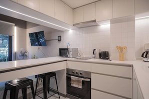 A modern kitchen with white cabinetry, a built-in oven, a microwave, a kettle, and a coffee maker. There are two black bar stools and a flat-screen TV mounted on the wall. - Study with a large window · Featured design (H06) (Providencia)