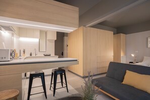 Cozinha moderna com ilha central, dois banquetes, um sofá cinza com almofada amarela e uma pequena planta.
