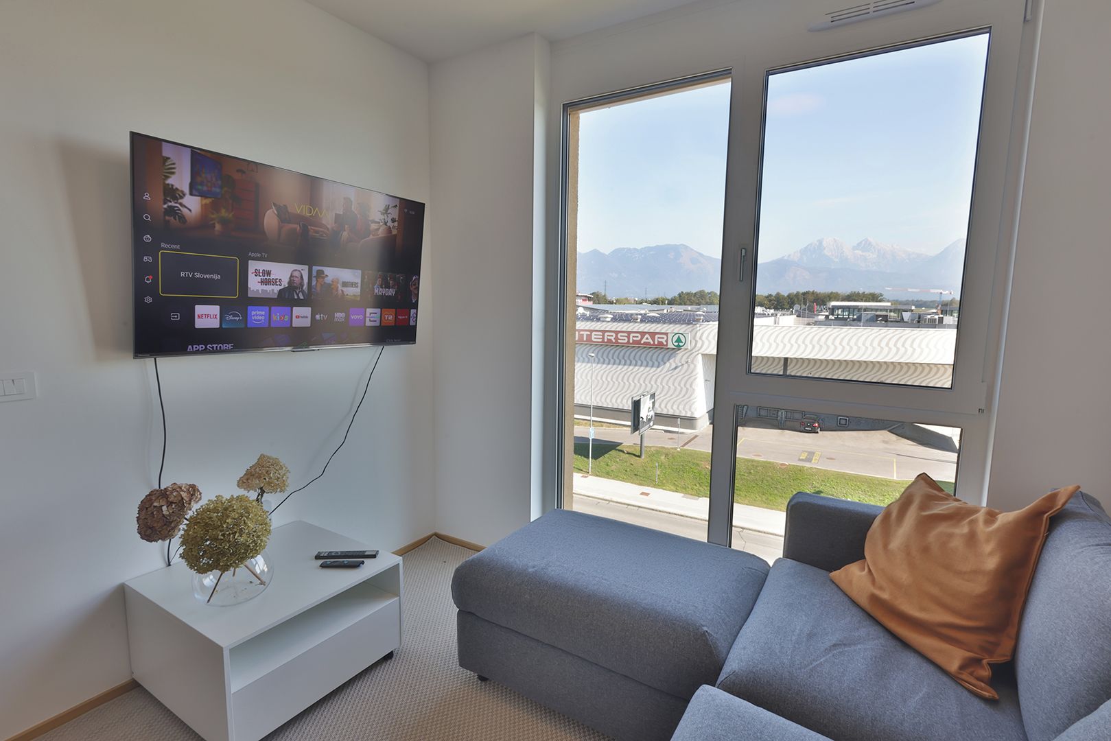 A modern living room with a flat-screen TV mounted on the wall, a white entertainment unit, a grey sofa with a brown cushion, and a large window with a view of an airport and mountains.