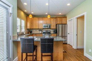A kitchen with wooden cabinets, a stainless steel refrigerator, a microwave, and a gas stove. There are two bar stools and pendant lights above the counter. - Spacious SLC Home | Hot Tub, Game Room, BBQ & Yard (Salt Lake City)