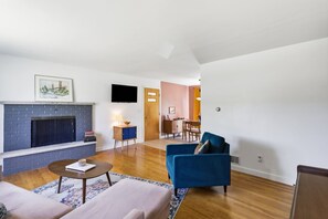 A living room with a fireplace, a blue armchair, and a dining area in the background. - Mid Century-Modern Retreat Great Allegheny Passage (Cresaptown, MD)