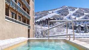 Ein Schwimmbecken mit Metallgeländer, ein Gebäude mit Balkonen und eine verschneite Berglandschaft im Hintergrund.