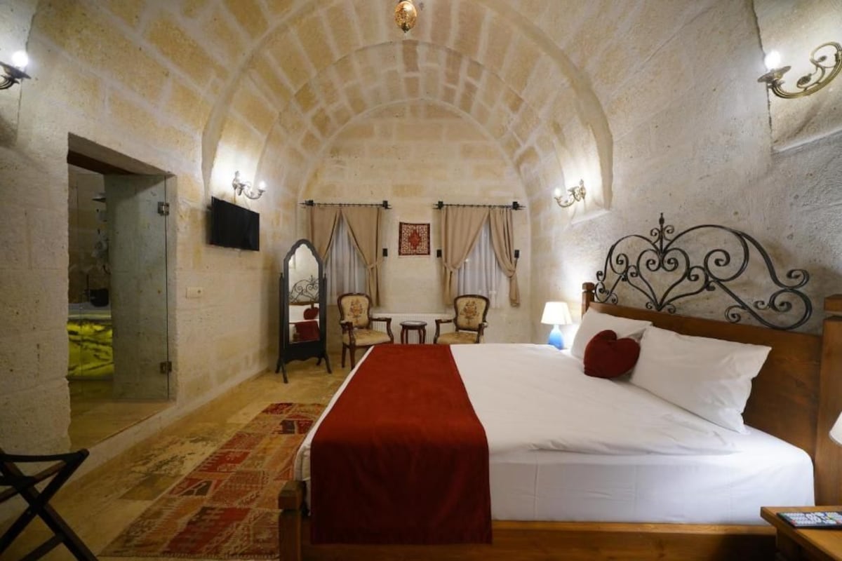 Habitació d'hotel amb un llit gran, una manta vermella, un panell de fusta al capçal, una taula petita amb cadires i un mirall a la paret.