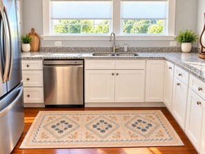 A kitchen with white cabinets, a stainless steel dishwasher, a granite countertop, a window with blinds, and a patterned rug. - The Biloxi Muse on Lameuse St. (Biloxi)