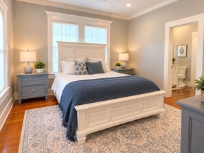 A bedroom with a large bed, two bedside tables, a nightstand, a window, and a door leading to a bathroom. - The Biloxi Muse on Lameuse St. (Biloxi)