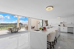 A modern kitchen with a large island, white cabinetry, and a view of a cityscape through large windows. - Luxury 2-Story Penthouse - Diamond Head & Ocean Views - Private Jacuzzi (Honolulu)