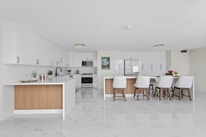 A modern kitchen with white cabinetry, a central island, and a dining area with a white table and chairs. - Luxury 2-Story Penthouse - Diamond Head & Ocean Views - Private Jacuzzi (Honolulu)