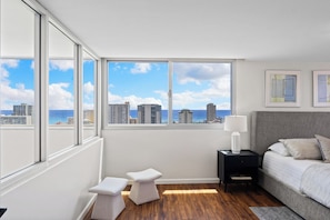 A modern bedroom with a large window, a bed, a bedside table, and a small stool. - Luxury 2-Story Penthouse - Diamond Head & Ocean Views - Private Jacuzzi (Honolulu)