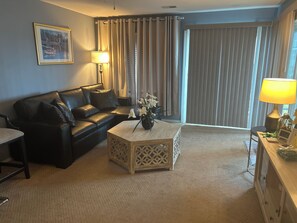 Ein Wohnzimmer mit einem schwarzen Ledersofa, einem Holztisch mit Blumenvase, einer Lampe und einem Bild an der Wand.