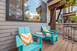 Two turquoise chairs with orange pillows on a wooden deck, a stone wall, and a large window reflecting trees. - Winter Park Escape -Luxury, Hot Tub, Pet Friendly (Winter Park)