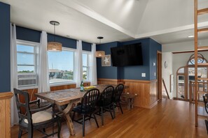 A dining area with a wooden table, chairs, and a television mounted on the wall. - Oceanside, 5 Story, w/Tiki Bar, 4th Flr HotTub, Panoramic view of Ocean & Sound! (Avon)