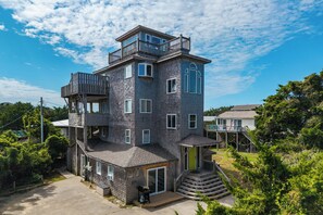 A multi-story house with a balcony on the upper level, a covered front porch, and a staircase leading to the entrance. - Oceanside, 5 Story, w/Tiki Bar, 4th Flr HotTub, Panoramic view of Ocean & Sound! (Avon)