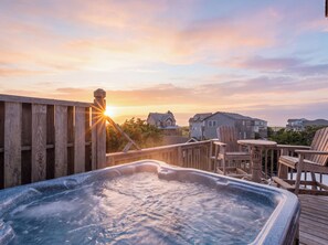 A wooden deck with a hot tub, outdoor furniture, and a sunset view of houses. - Oceanside, 5 Story, w/Tiki Bar, 4th Flr HotTub, Panoramic view of Ocean & Sound! (Avon)