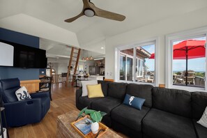 A modern living room with a dark sofa, a coffee table with a magazine and a plant, a TV mounted on the wall, and a view of the kitchen and outdoor patio through large windows. - Oceanside, 5 Story, w/Tiki Bar, 4th Flr HotTub, Panoramic view of Ocean & Sound! (Avon)