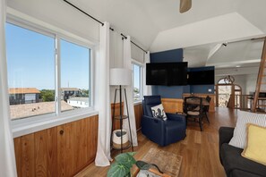 A spacious living area with a large window, a flat-screen TV, a blue sofa, and a dining table. - Oceanside, 5 Story, w/Tiki Bar, 4th Flr HotTub, Panoramic view of Ocean & Sound! (Avon)