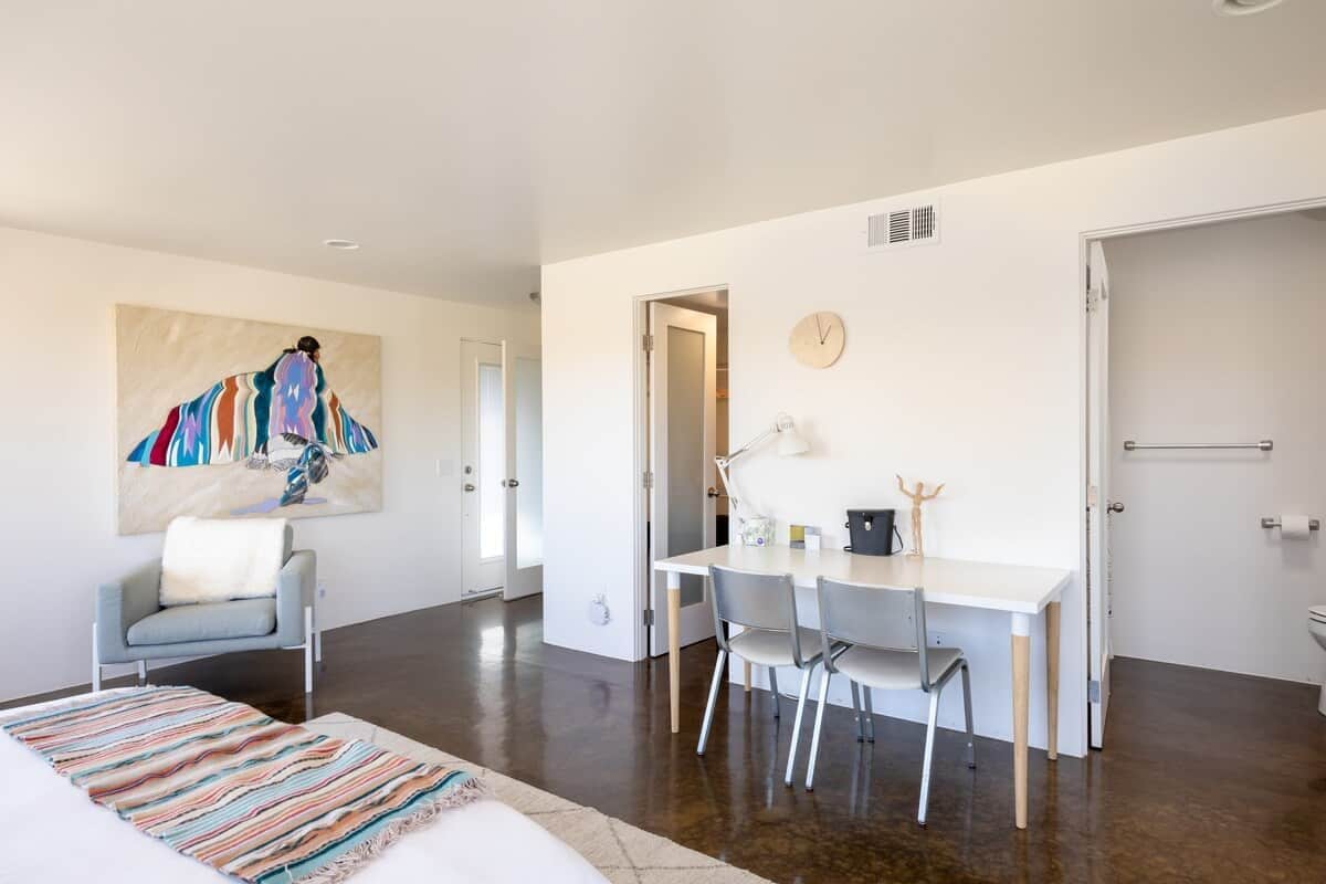 A modern room with a bed, a desk, and a chair. There's a painting on the wall and a clock.