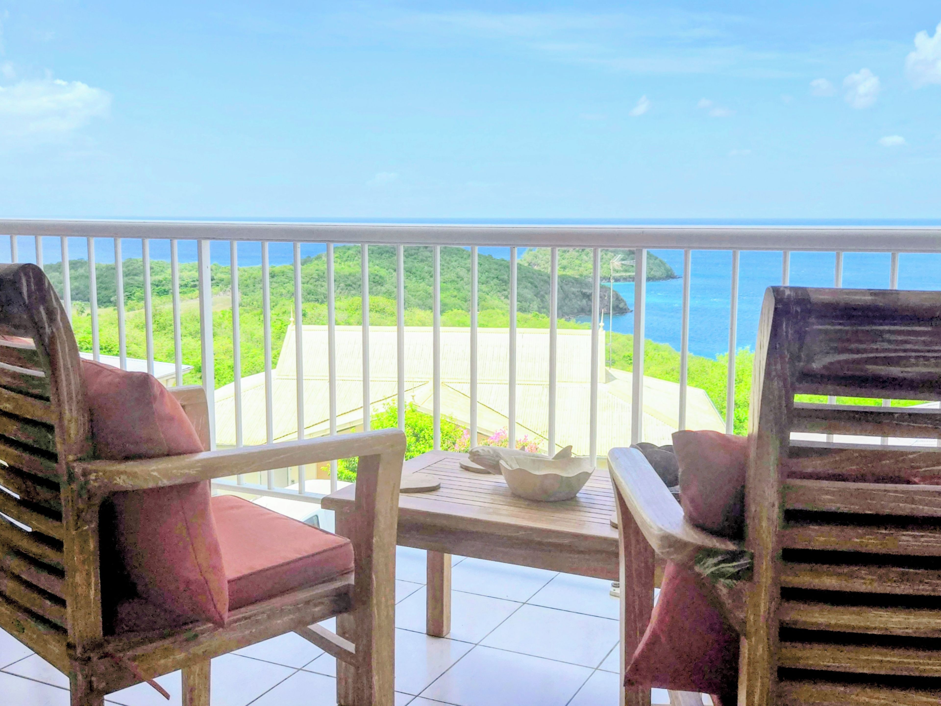 A balcony with two wooden chairs and a table, overlooking a coastal landscape with greenery and the sea.