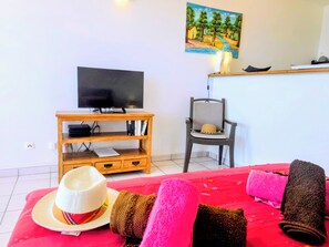 Ein Zimmer mit Fernseher auf Holzfuß, einem Sessel, einem Bett mit rosa Bettwäsche und einem Gemälde an der Wand.