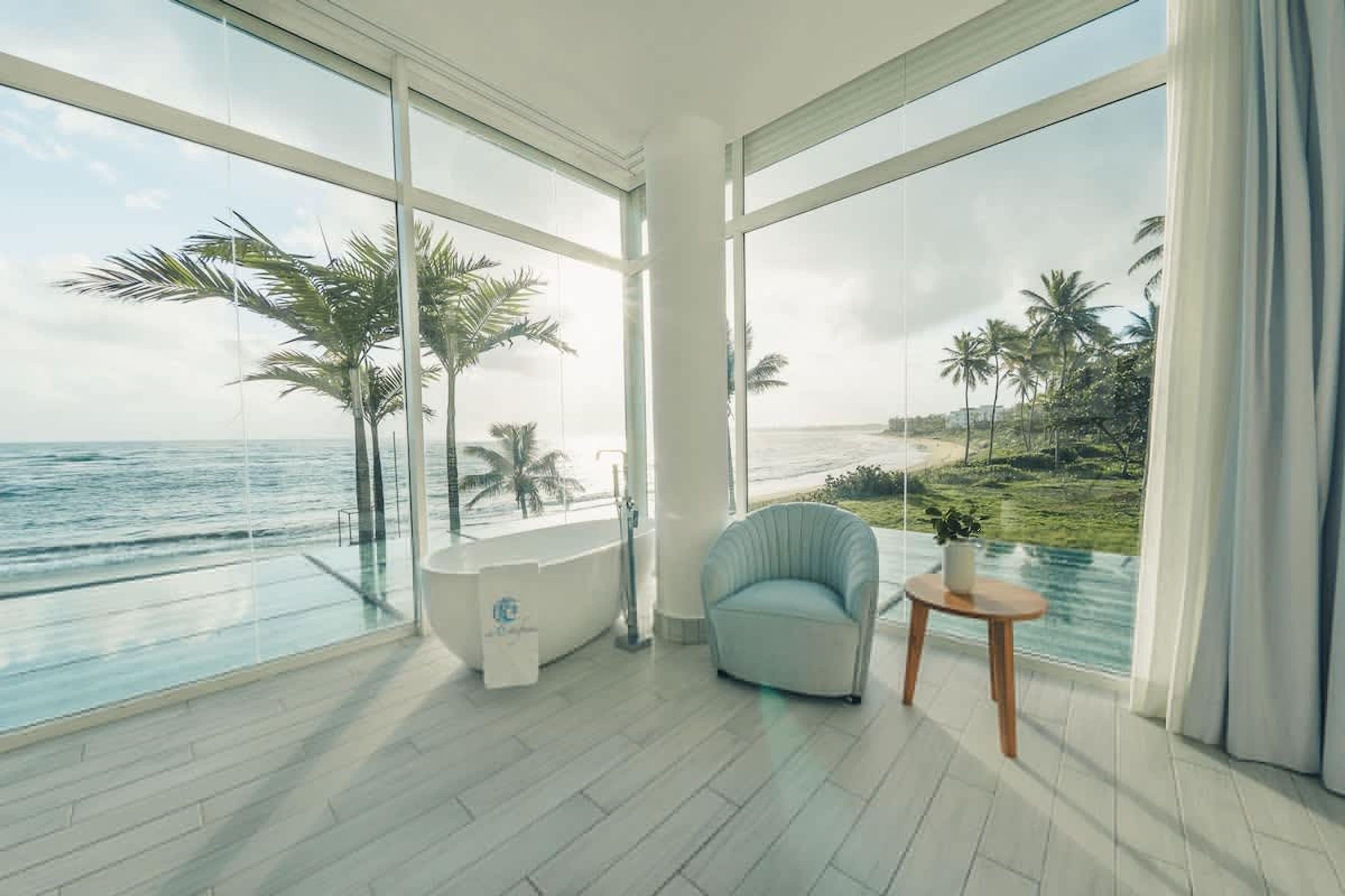 A modern bathroom with a bathtub, a chair, and a small table, overlooking a beach with palm trees and the ocean.