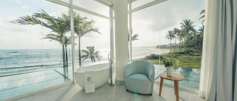 A modern bathroom with a bathtub, a chair, and a small table, overlooking a beach with palm trees and the ocean.