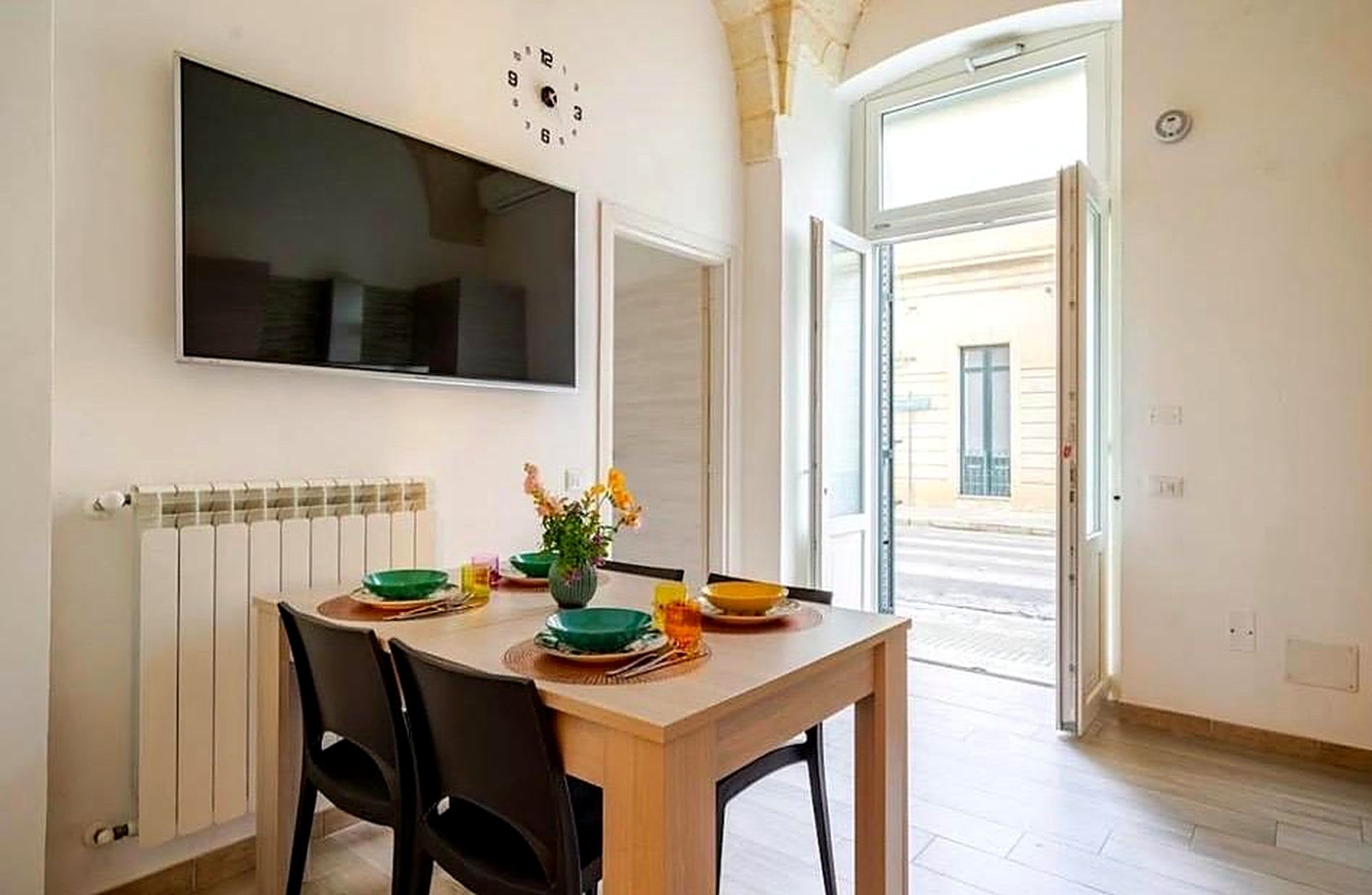 A dining area with a table set for two, a wall-mounted clock, and a radiator.