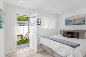 A bedroom with a bed, a window with curtains, a painting on the wall, and a door leading to an outdoor area with a table and chair. - Two bedroom house in quaint Dania Beach on a canal and next to airport (Dania Beach)