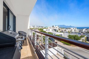 A balcony with a grill and seating, overlooking a residential area with buildings and a clear sky. - Azure 119 - Two Bedroom Apartment, Sleeps 4 (Cape Town)