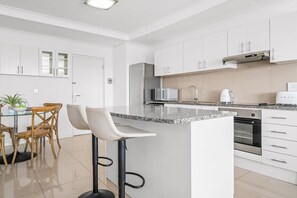 Cozinha moderna com armários brancos, bancada de granito, forno embutido e área de refeições com banquetas.