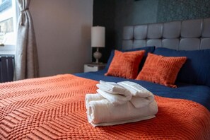 Ein ordentlich bezogenes Bett mit einem gefalteten weißen Handtuch, einer blauen und orangefarbenen Bettdecke und zwei Kissen.