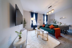Ein modernes Wohnzimmer mit einer Couch, zwei Beistelltischen, einem Flachbildfernseher und einem Essbereich mit Tisch und Stühlen.