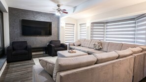 A modern living room with a sectional sofa, two armchairs, a flat-screen TV mounted on a textured wall, and a ceiling fan. - Brand New - Private Renovated Estate - Secluded! (Brampton)